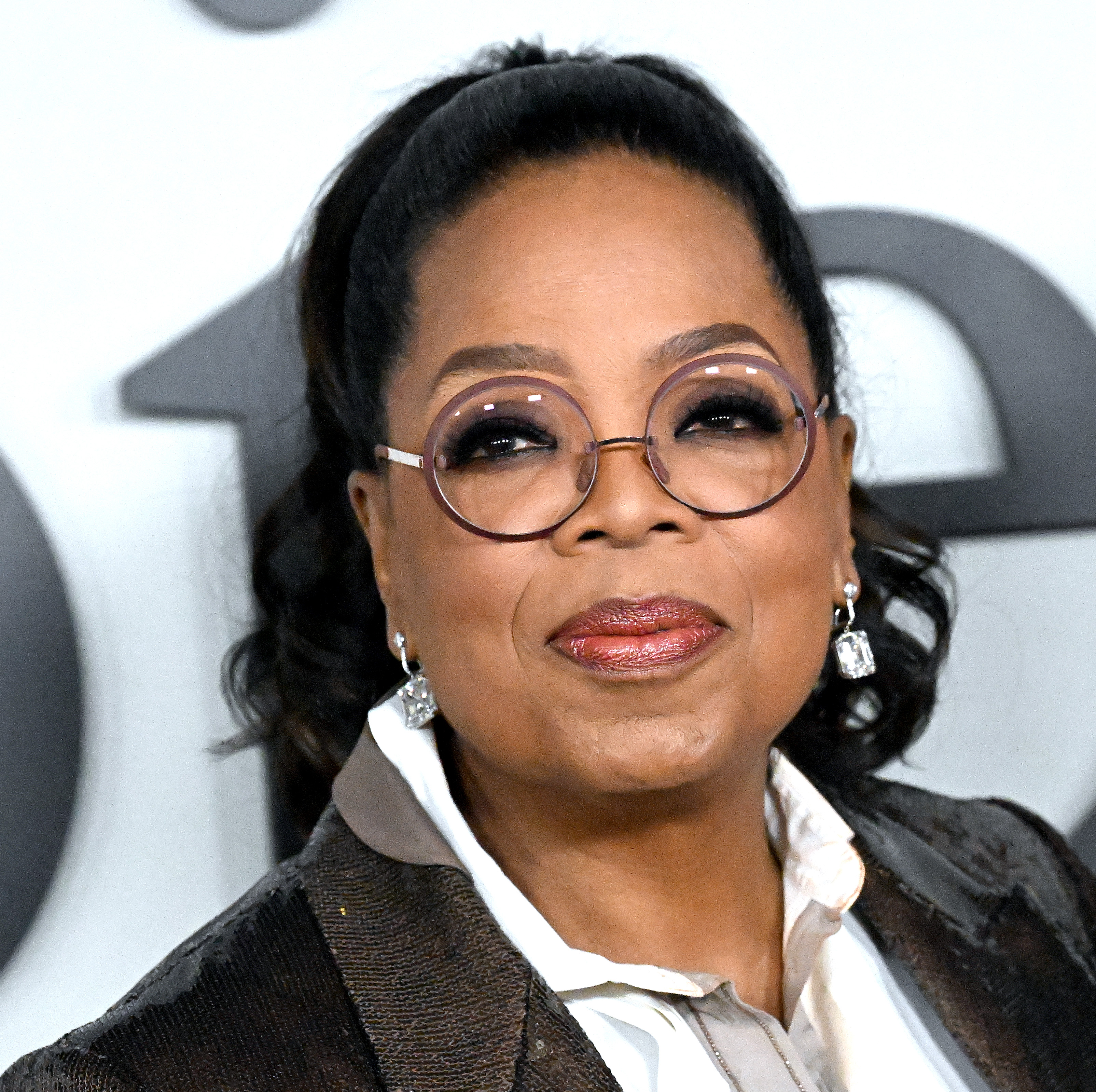 Oprah Winfrey wearing glasses and a black jacket, smiling at the camera.