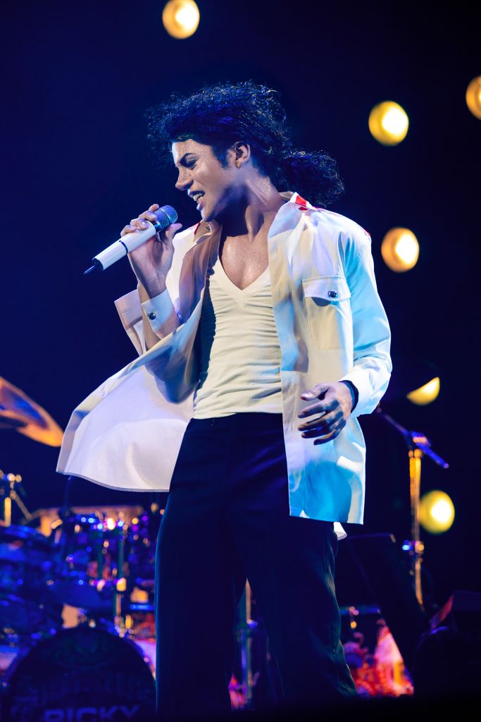 A performer with curly dark hair singing into a microphone on stage, wearing a white jacket and black pants, with a blue and white background and spotlights.