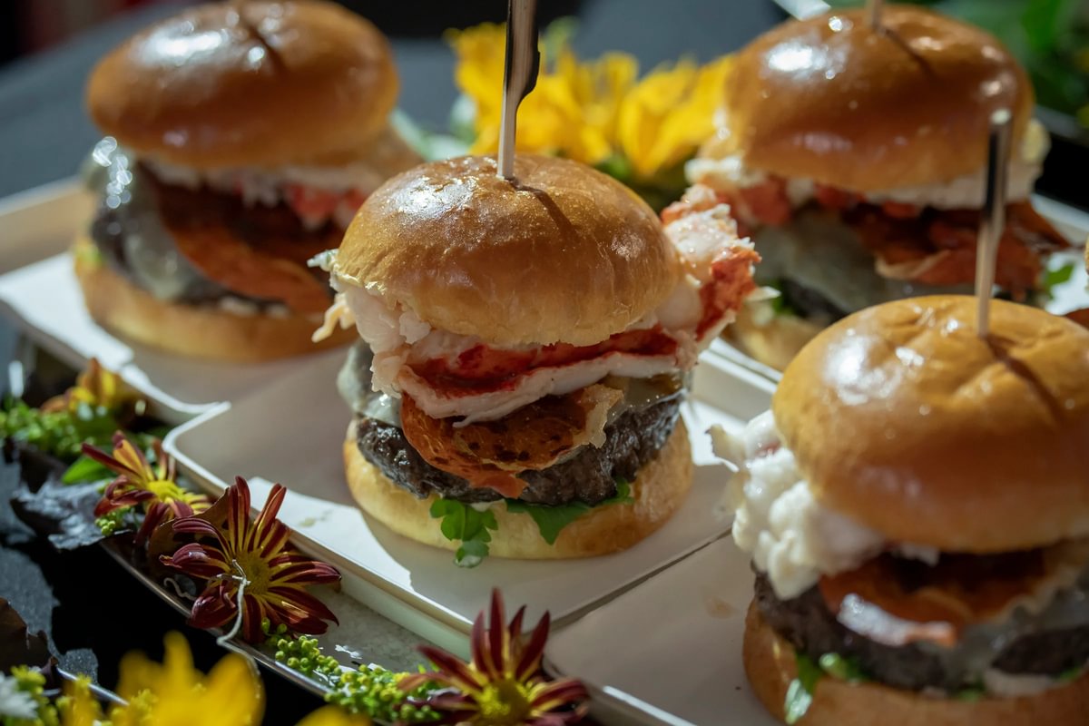 Assorted hamburger sliders with lettuce, tomato, and other toppings, arranged on a plate with yellow and red flowers.