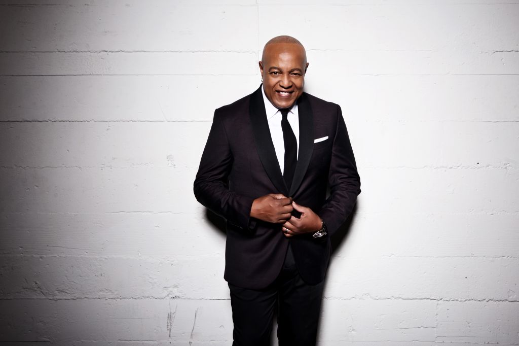 A smiling middle-aged Black man in a dark suit standing against a plain white wall.