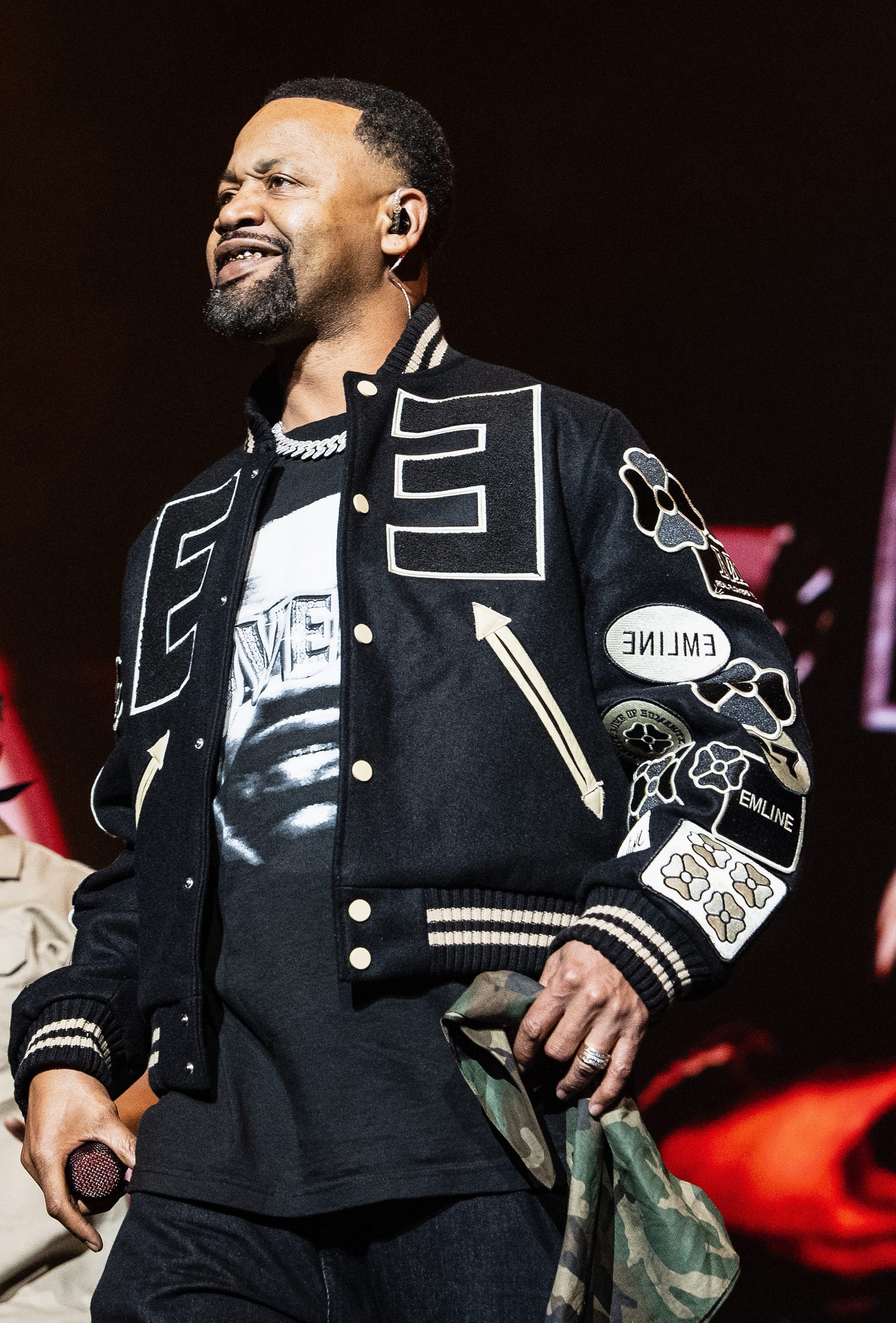 A man in a black jacket with graphic designs performs on stage, with another man in a beige jacket visible in the background.