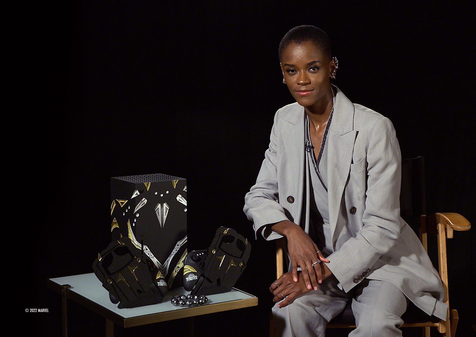 A woman in a gray suit sitting in a chair, with a Black Panther: Wakanda Forever movie poster and merchandise visible.