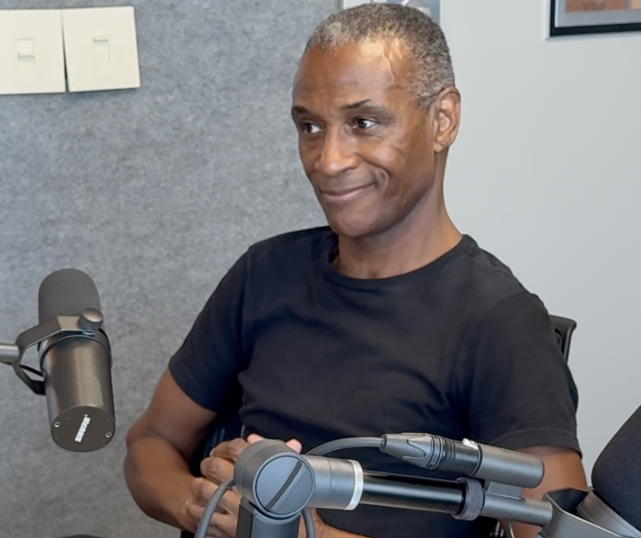 A smiling middle-aged Black man sitting in a radio studio, holding two microphones and surrounded by audio equipment.